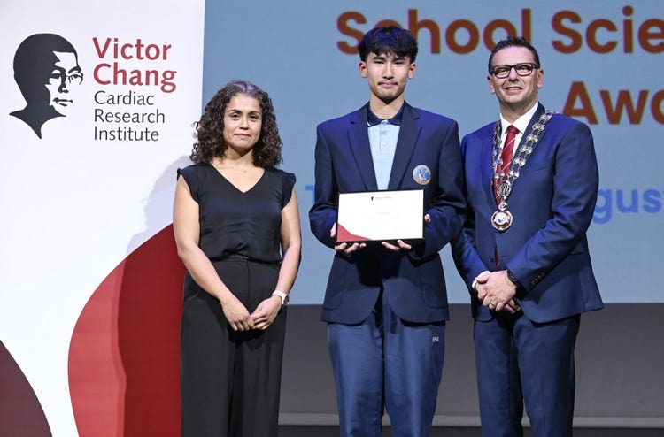 A student standing on stage receiving the Victor Chang Award next to another female and male member of the community.